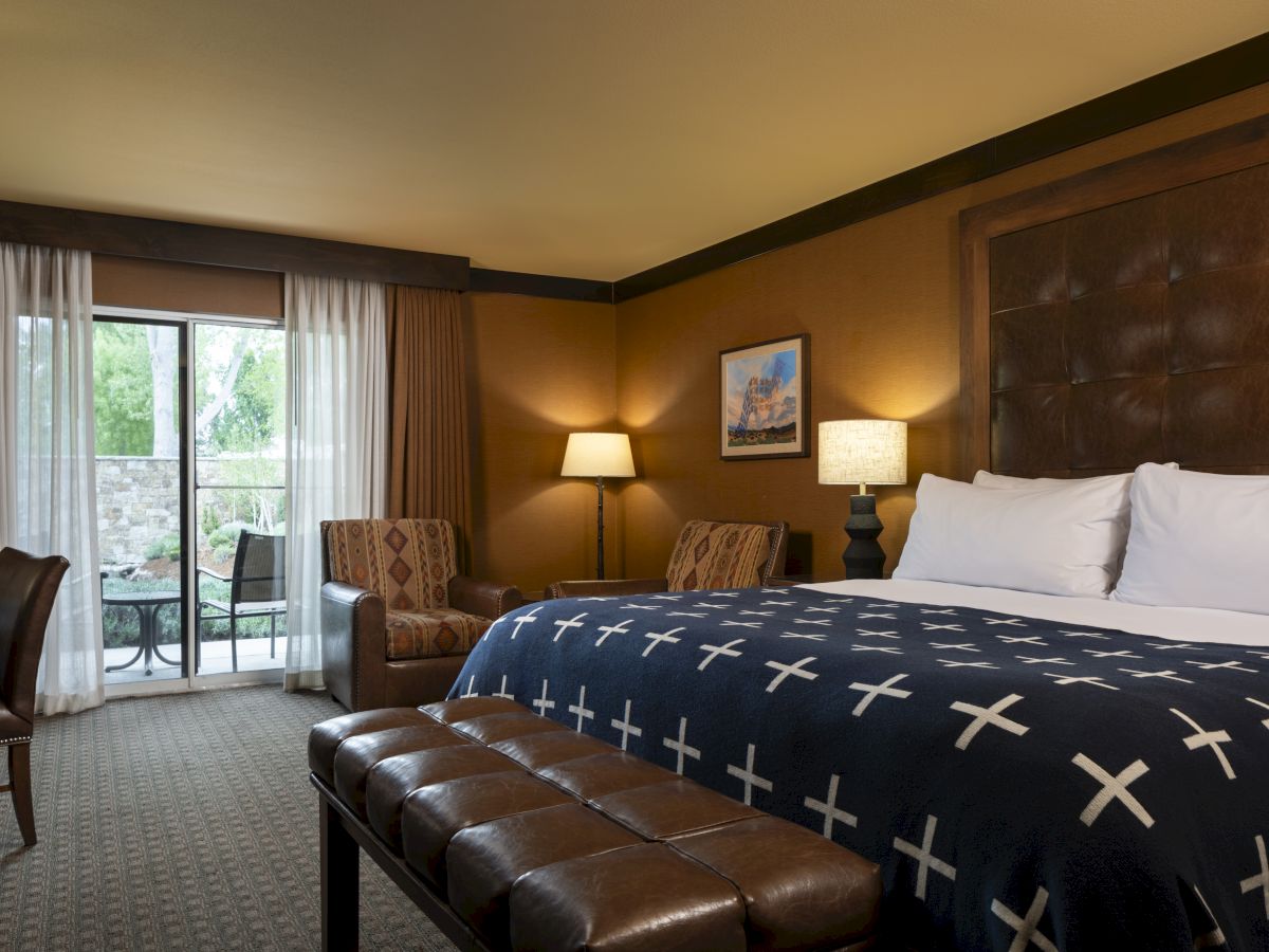A hotel room with a king bed, brown headboard, navy bedspread with white crosses, two bedside lamps, a bench at foot, seating area, and a balcony door with sheer curtains.