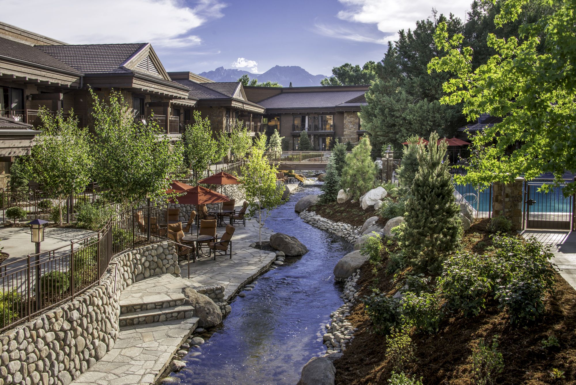 A scenic resort with a stone path, a winding river, lush trees, landscaped rocks, and wooden buildings lining the water, mountains in the background.