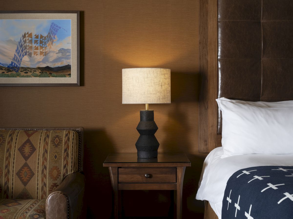 A cozy hotel room scene with a striped sofa, a wooden nightstand, a geometric black lamp, a bed with white sheets and a dark blue cross pattern duvet, and framed art on a brown wall, all lit softly.