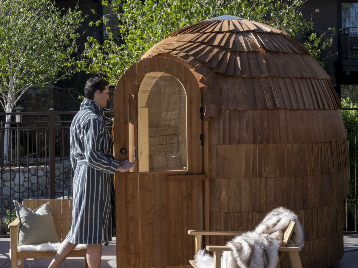 A wooden dome sauna-like pod with a small arched door, sitting outdoors; a person in striped clothing stands nearby, a dog lounging on a chair.