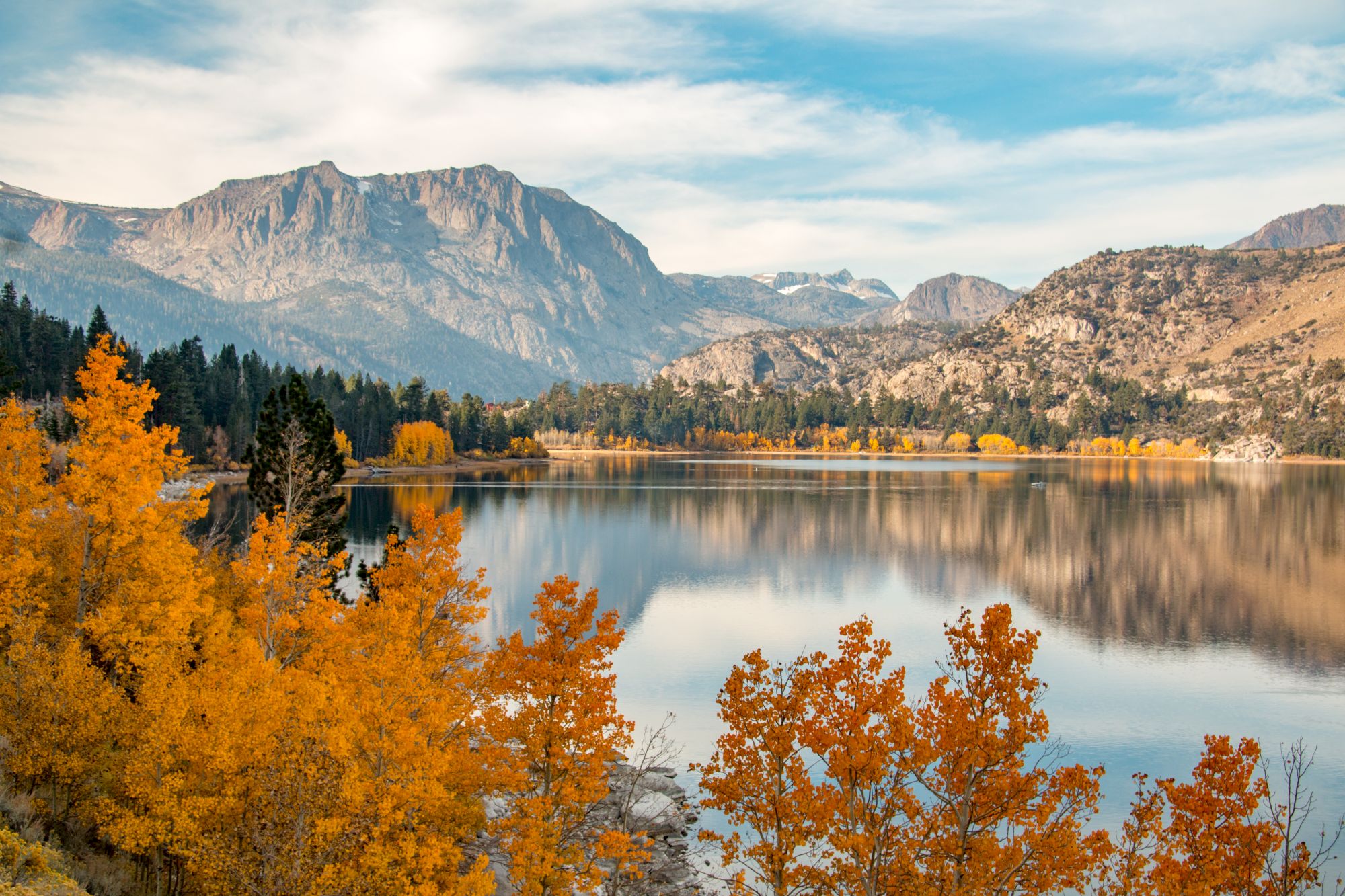 A serene lake reflects mountains and a sky, framed by vibrant autumn trees in orange and yellow hues under a partly cloudy sky.