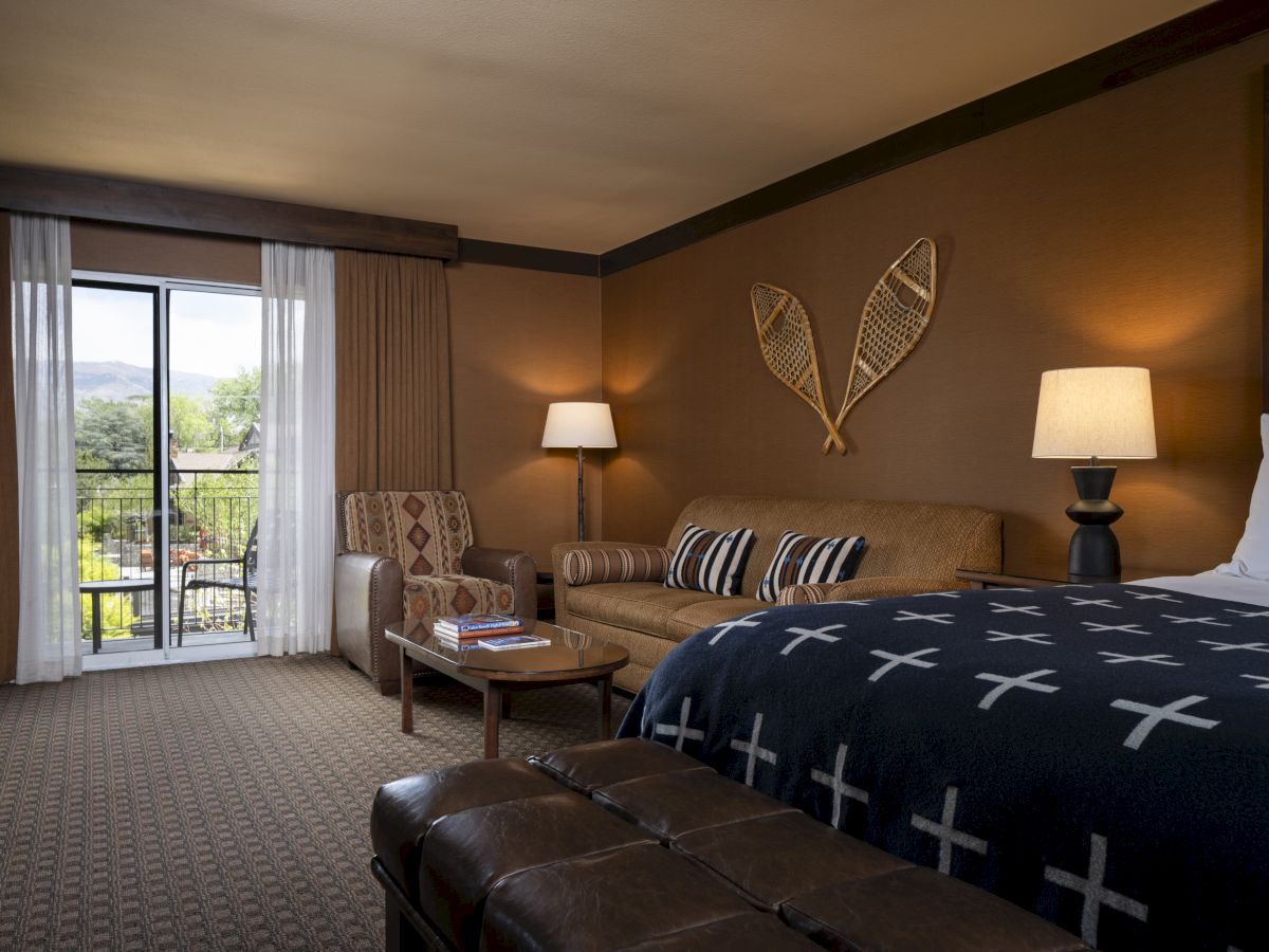 A cozy hotel room with a large bed, loveseat, two lamps, and a sliding door to a balcony overlooking greenery.