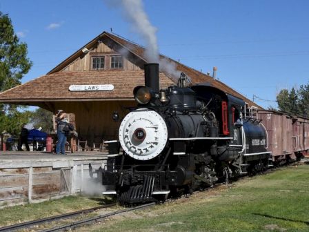 A vintage steam locomotive pulls into a rural station with a wooden depot, onlookers nearby and cars along the tracks.