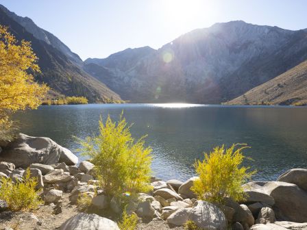 A tranquil mountain lake surrounded by rocky shore, golden-yellow shrubs, and clear water with sunlit peaks in the background.