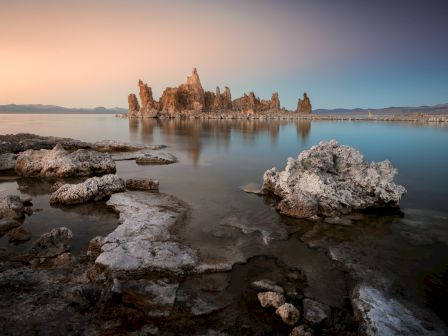 Rocky shoreline with calm water and dramatic limestone pillars rising from a distant island at sunset, soft pastel sky reflecting on the surface.