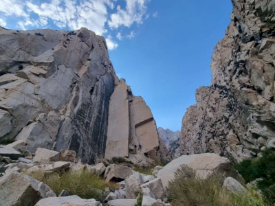 Rugged canyon with tall rock walls, boulders on the ground, sparse vegetation, and a clear blue sky above.