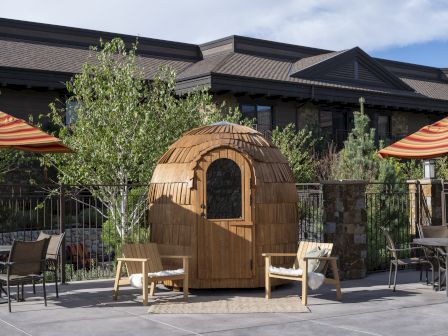 A wooden, rounded gazebo-like hut at a patio with chairs and orange umbrellas, set against a modern building background.