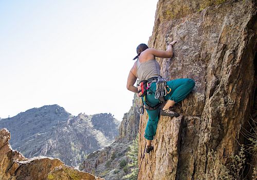 Two climbers scale a steep rock face on a rugged mountainside, with one climber higher up gripping the rock as teammates below assist.
