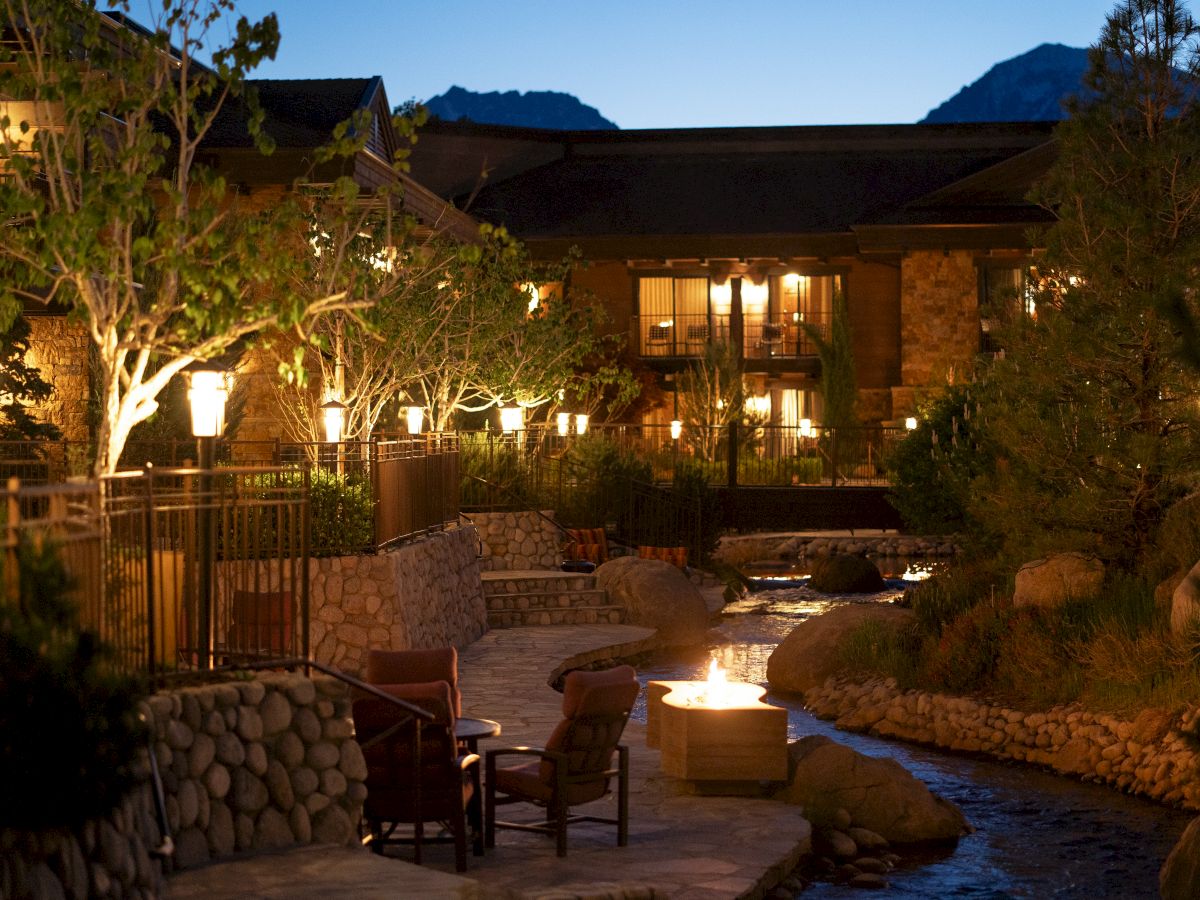 A cozy outdoor resort at dusk with a lit courtyard, stone paths, seating around a glowing fire pit, and a tranquil stream winding through.