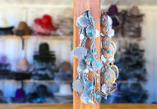 Shoe accessories hang on a wooden peg in a shop, with denim-fringed pieces and turquoise beads, while hats line the shelves in the background.