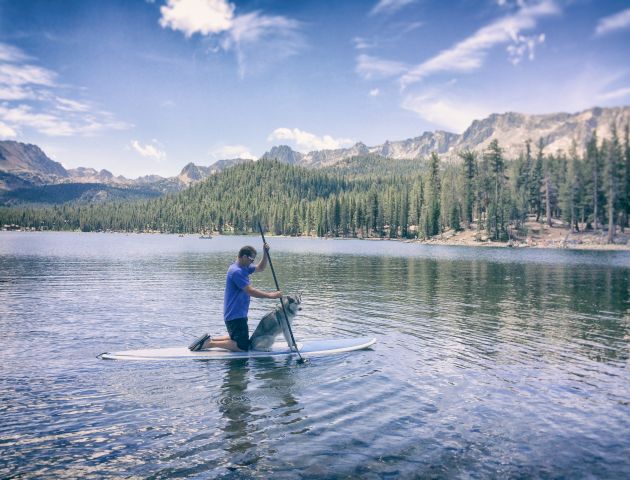 A person stands on a paddleboard, paddling on a calm lake surrounded by pine trees and mountains under a blue, sunny sky.
