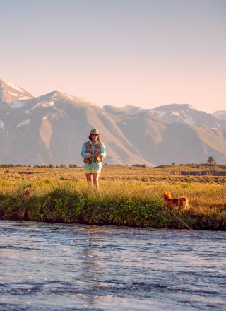 Two people fishing near a river with mountains in the background, a dog nearby, golden-hour light, wide open grassy bank.