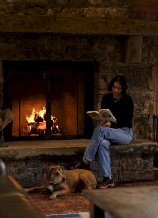 A person sitting by a stone fireplace reading a book while a dog rests on the floor beside them.