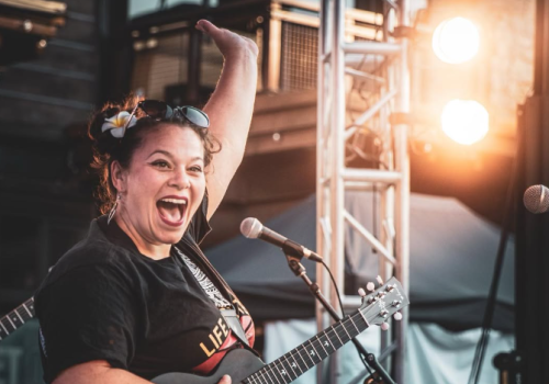A joyful female musician singing and playing a ukulele on stage, with bright lights and a cheering vibe.