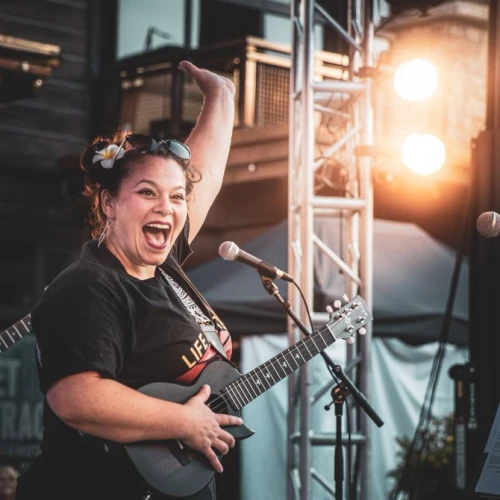 A joyful female musician singing and playing a ukulele on stage, with bright lights and a cheering vibe.