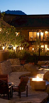 A warmly lit outdoor retreat with stone paths, a fire pit, seating by a small stream, and a lit building in the background under a twilight sky.