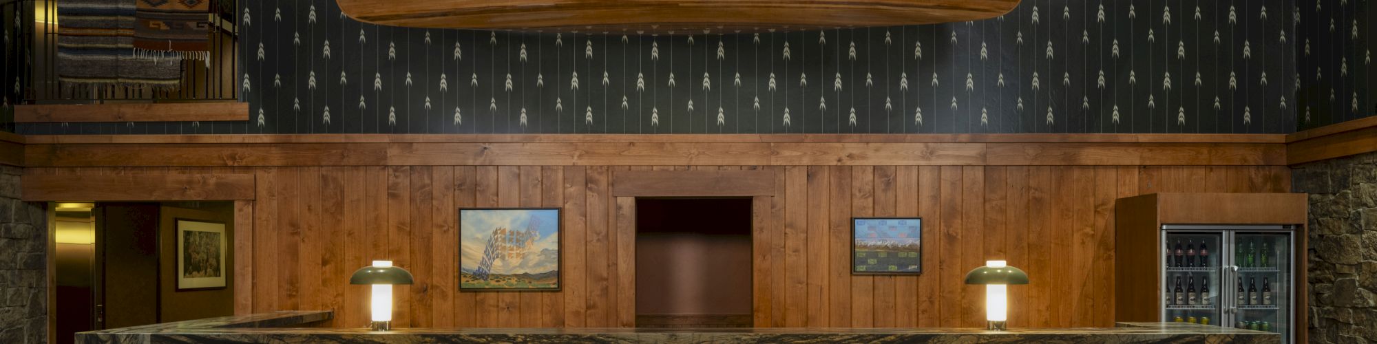 A rustic hotel lobby with a wooden front desk, warm lighting, dark patterned wall, and a curved wooden sculpture hanging above the desk.