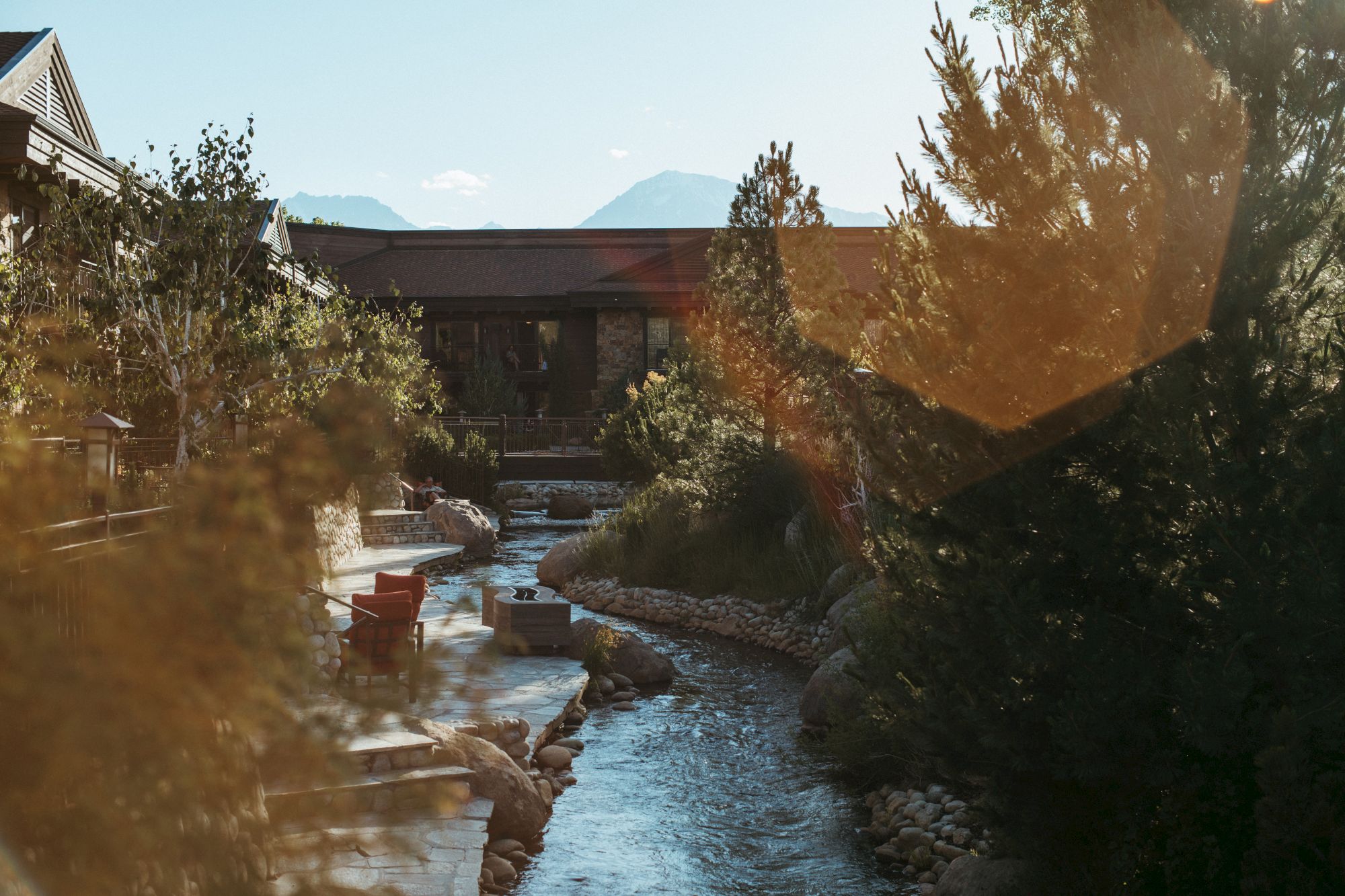 A tranquil riverside garden with a rocky stream, wooden seating, and lush trees, bathed in warm sunlight by a calm, winding waterway.