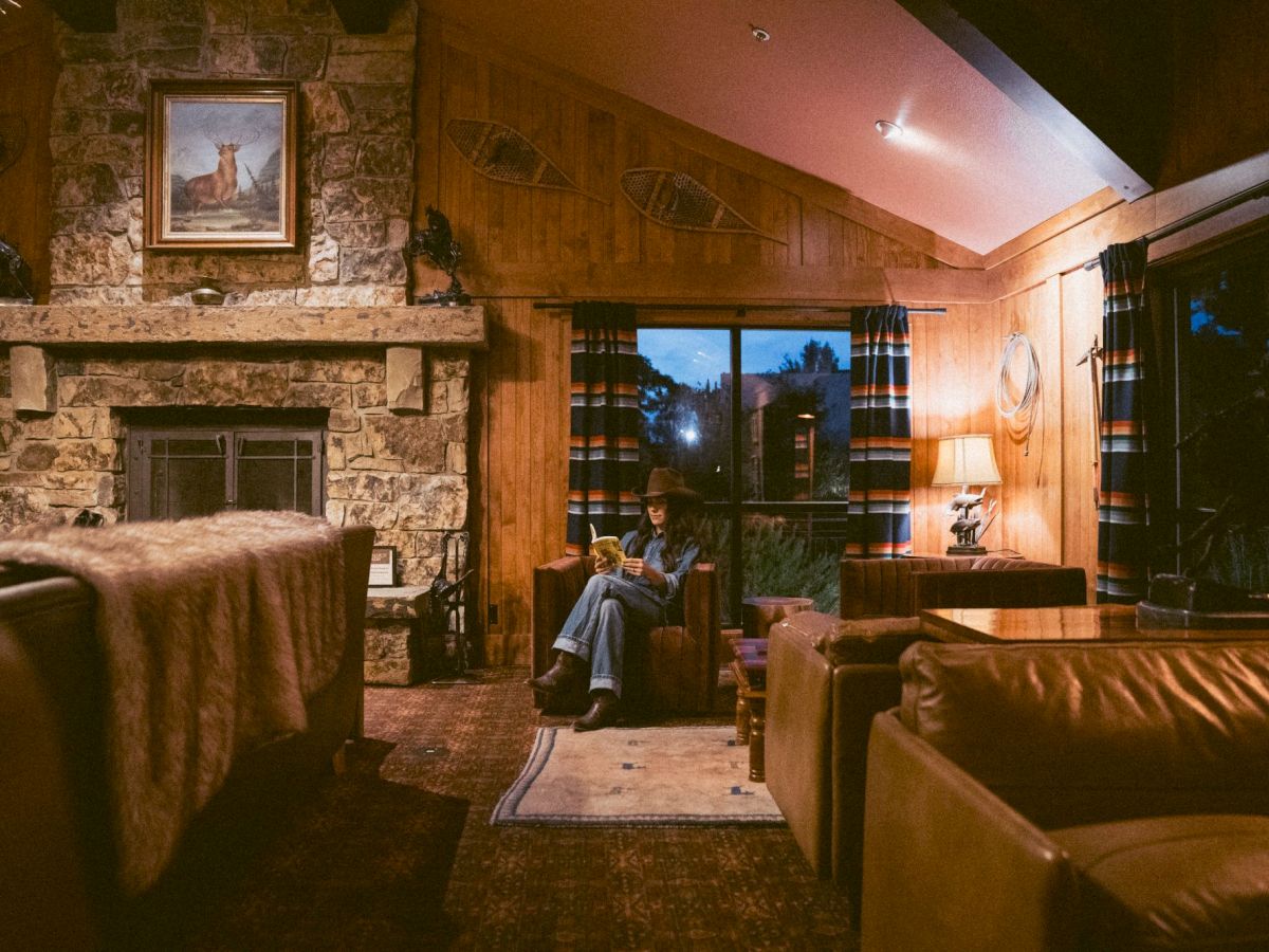 Cozy rustic living room with a stone fireplace, wooden beams, and leather sofas; a person sits near a window in warm ambient light.