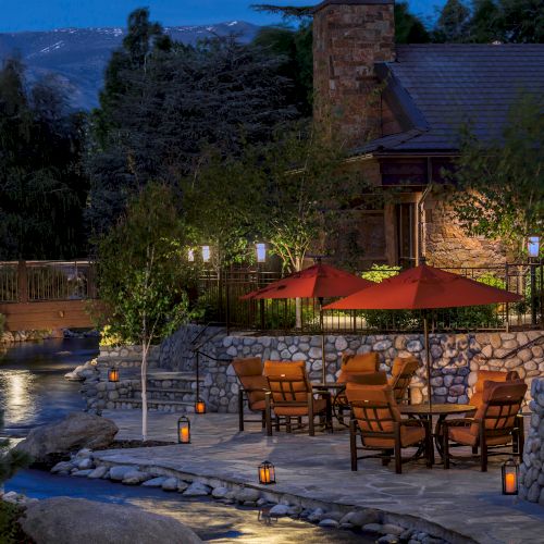 An outdoor stone-terrace dining area at dusk, with red umbrellas, lanterns along a stone path, trees, and a cozy lodge-like building in the background.