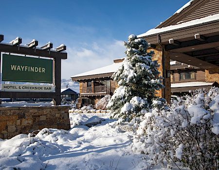 Snow-covered hotel entrance with a “WAYFINDER” sign reading “Hotel & Creekside Bar” and buildings and trees capped in snow.