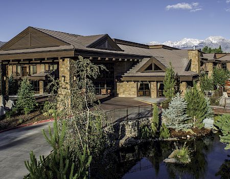 A mountain lodge/hotel with stone-and-wood architecture, landscaped gardens, a pond, and snowy peaks in the background.
