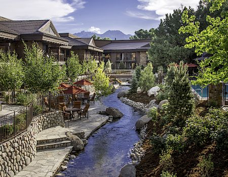 A landscaped courtyard with a small stream, patio seating and umbrellas, surrounded by lodge buildings and bright green trees.