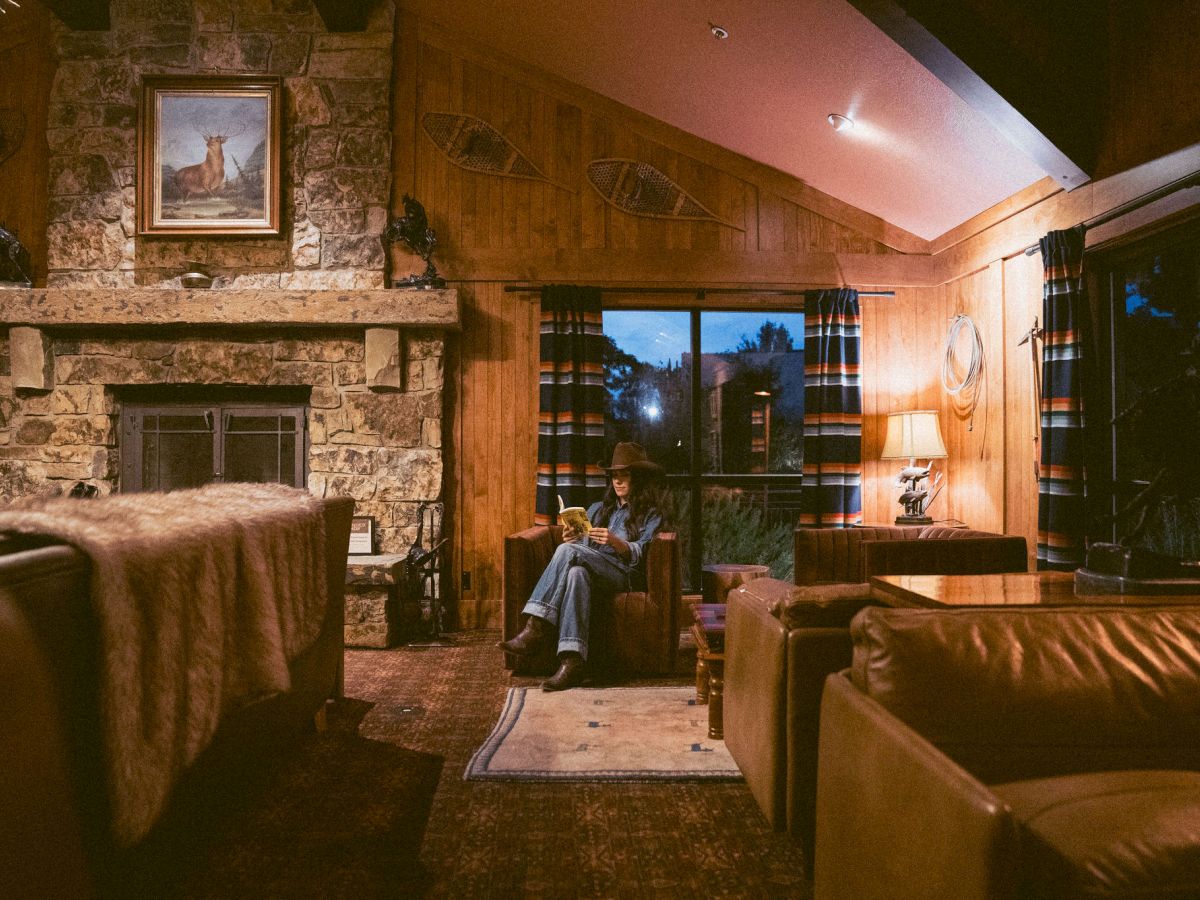 A cozy living room with a stone fireplace, deer painting, curtains, and a person sitting and reading on a couch.