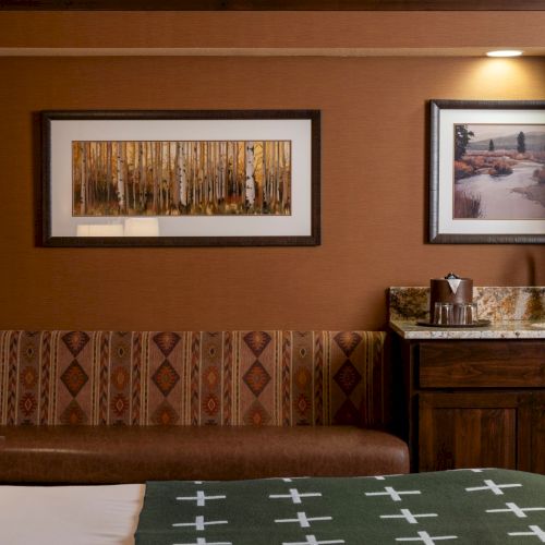 A cozy hotel room corner with a bed, patterned cushions, two landscape artworks, and a small vanity sink with coffee items.