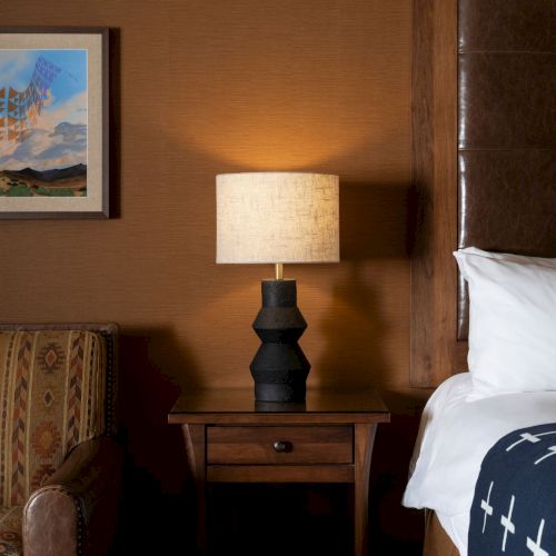 A cozy bedroom corner with a bed, a table lamp, framed artwork, and patterned textiles on a chair or ottoman.