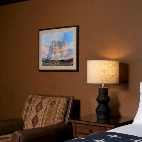 A cozy hotel room corner with a framed artwork, a lit table lamp, a patterned chair, and a bed with a blue blanket.