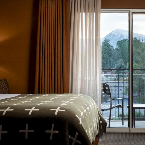 A cozy hotel bedroom with a lamp, bed, and curtains, looking out onto a balcony with distant mountains beyond.