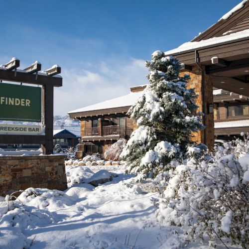 Snowy lodge entrance with a Wayfinder sign for Hotel & Creekside Bar and pine trees dusted with snow under a blue sky.