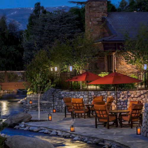 A cozy outdoor patio at dusk with wooden chairs, red umbrellas, lanterns, a stone wall, and a lodge overlooking mountains.