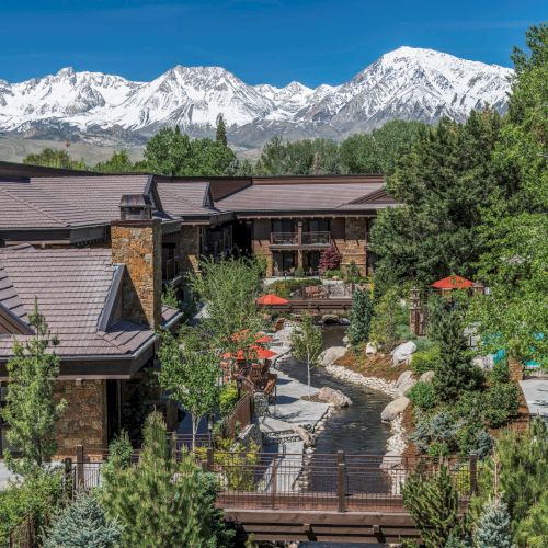 A mountain resort with stone cabins, red umbrellas, a small stream, and snowy peaks under a bright blue sky.