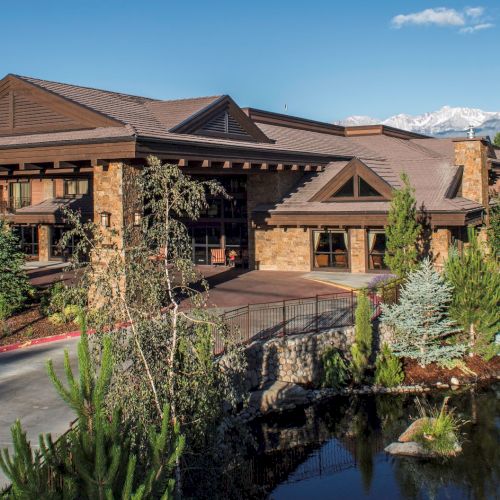 A stone-and-wood lodge with a landscaped courtyard and pond, set against snowy mountains under a blue sky.