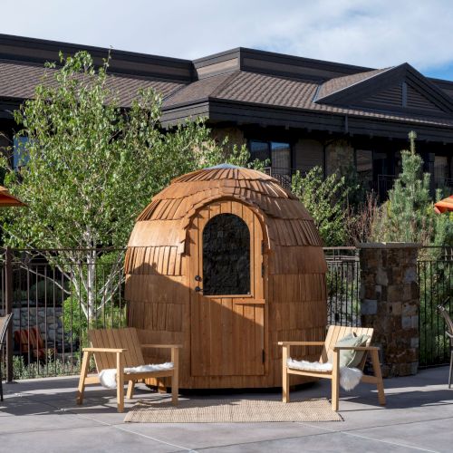 A backyard patio with two striped umbrellas, lounge chairs, and a small wooden dome playhouse in front of a modern home.