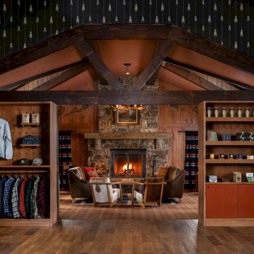 Rustic lodge interior with a roaring fireplace, wooden shelves, plaid blankets, and armchairs arranged in front of the fire.