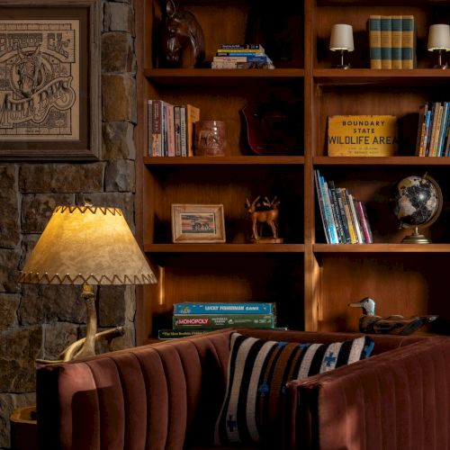 A rustic living room shelf display with books, lamps, framed art, and animal figurines (deer head and antlers) beside a couch.