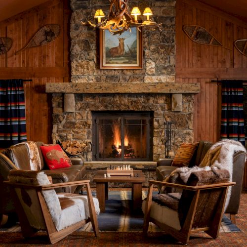 A cozy log cabin lounge with a roaring fireplace, deer artwork, antler chandelier, leather chairs, and a chessboard on tables.