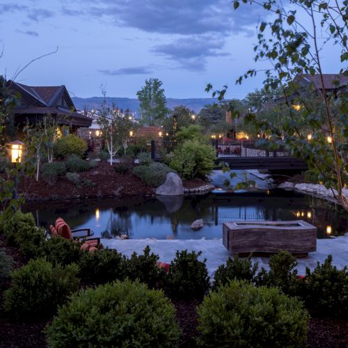 A dusk-lit backyard with a small pond, stone paths, warm lights, trees, and a cozy house overlooking distant hills.