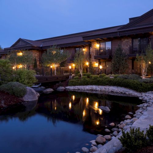 A dusk view of a luxury lodge with stone walls, glowing balconies, and landscaped gardens beside a calm reflecting pond.