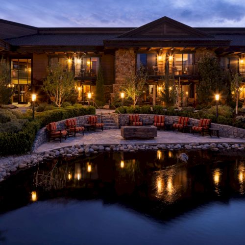 A warmly lit luxury home at dusk with a patio fire pit, striped lounge chairs, and a calm pond reflecting the lights.