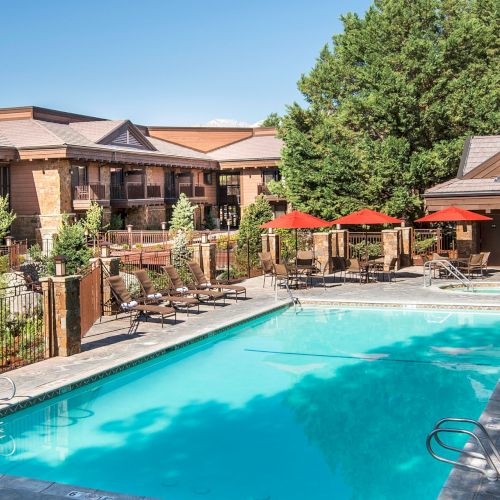 An outdoor resort pool with lounge chairs, red umbrellas, and a brown building surrounded by green trees under a blue sky.