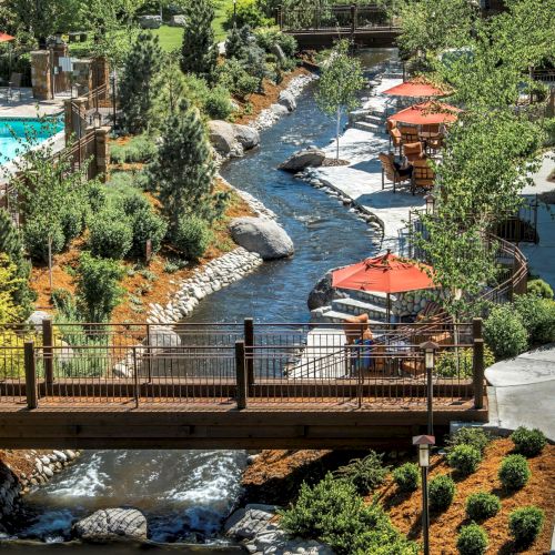 A lush resort courtyard with a turquoise pool, flowing stream, red patio umbrellas, and outdoor seating amid trees and stone paths.