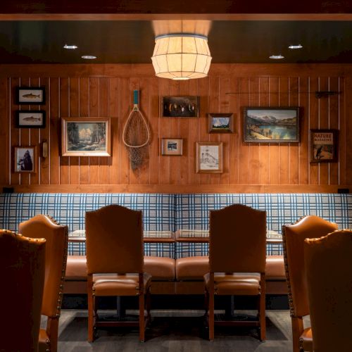 A cozy lodge room with stone walls, wood paneling, framed nature photos, warm chandelier light, and leather chairs and sofa.