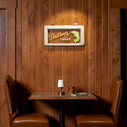 A cozy corner with wooden paneling, two leather chairs, and a small table featuring a lamp and mug beneath a &ldquo;Webb&rsquo;s&rdquo; trout sign.