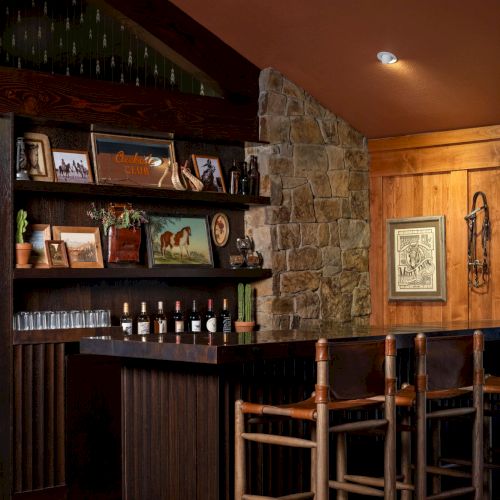 A cozy bar area with wood shelves, bottles and glassware, a stone fireplace wall, framed d&eacute;cor, and wooden stools in warm lighting.