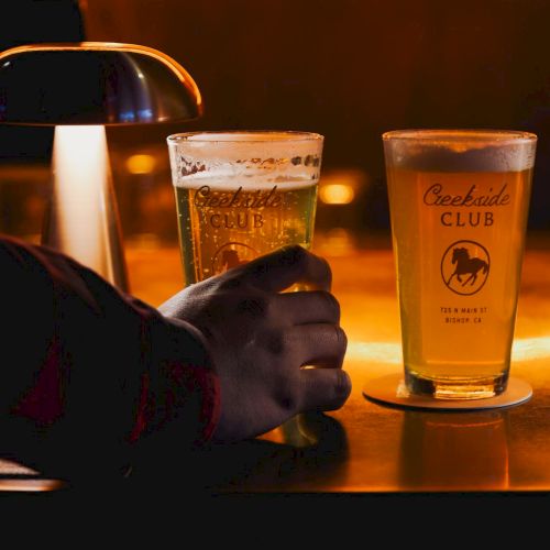 A hand clinks two tall beer glasses labeled &ldquo;Creekside Club&rdquo; at a warm bar, with frothy light amber drinks.