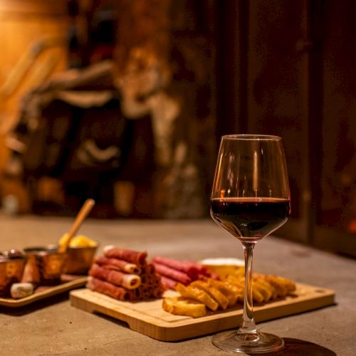 A glass of red wine rests on a board with bread, crackers, and cured meats, in warm candlelit blur behind.